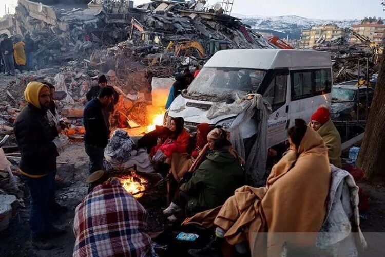 Orang-orang duduk di sekitar api di samping puing-puing dan kerusakan di dekat lokasi bangunan yang runtuh setelah gempa bumi, di Kahramanmaras, Turki, (08/02/2023). (Foto: Ant/Reuters/Suhaib Salem/pri).